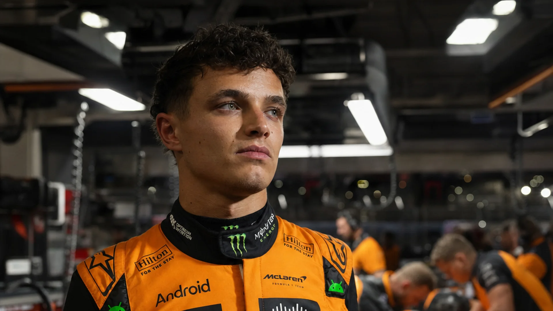 Lando Norris in McLaren-pak in de donkere garage in Monaco terwijl monteurs achter hem werken