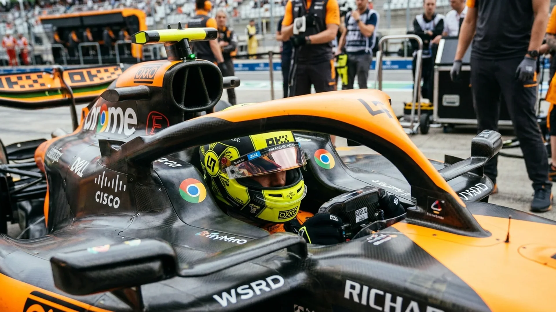 Gehelmde McLaren-coureur met neergeklapt vizier in de cockpit van de papaya-zwarte McLaren op de pitstraat