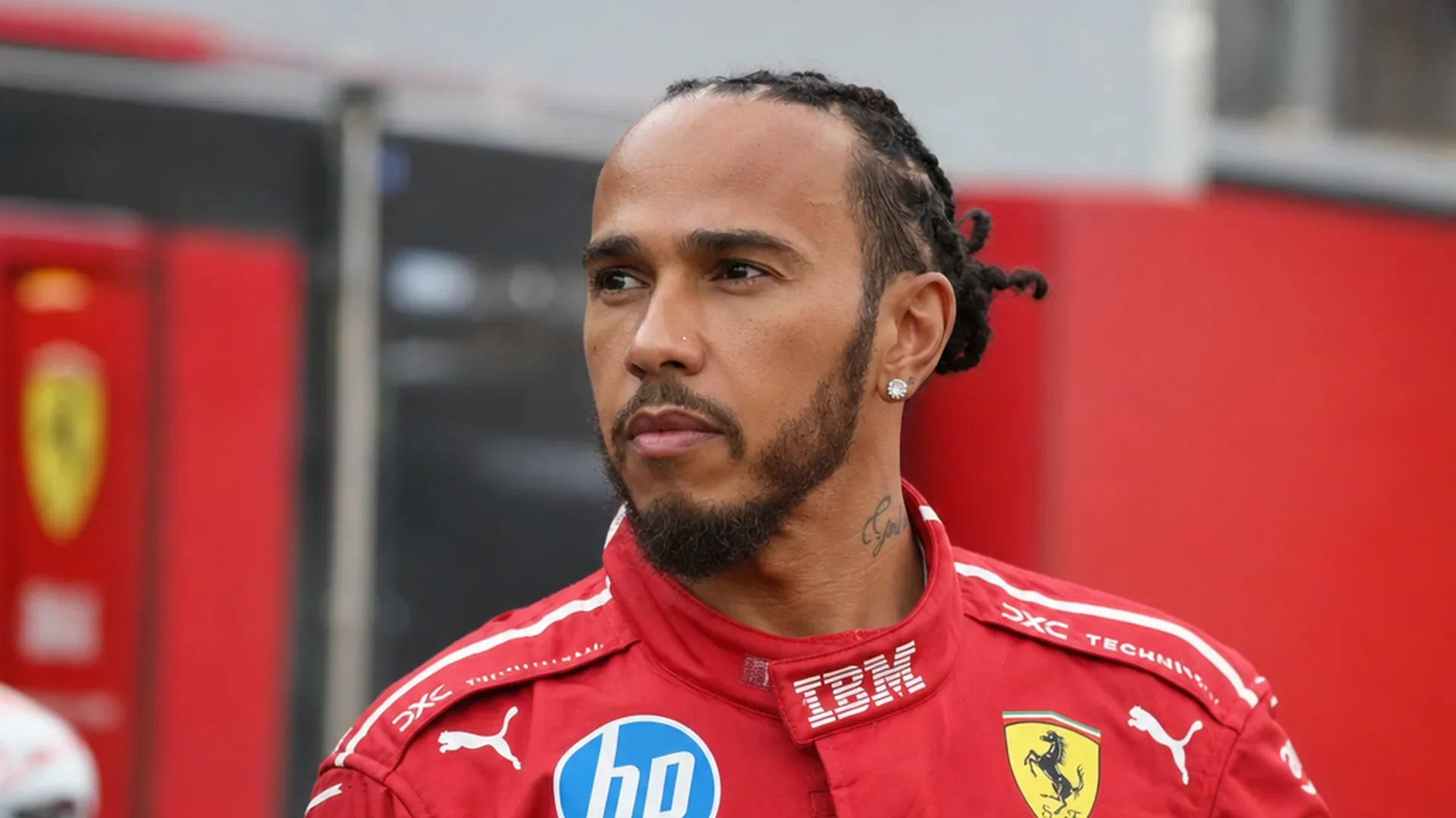Lewis Hamilton in het rode Ferrari-pak kijkt geconcentreerd voor zich uit in de paddock